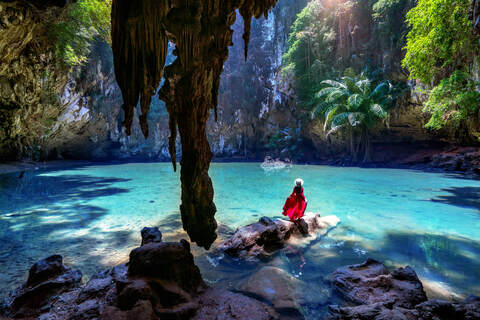 Woman in red dress looking at beautiful blue lagoon with stalactite, palm trees and greenery at princess lagoon at railay, krabi in thailand